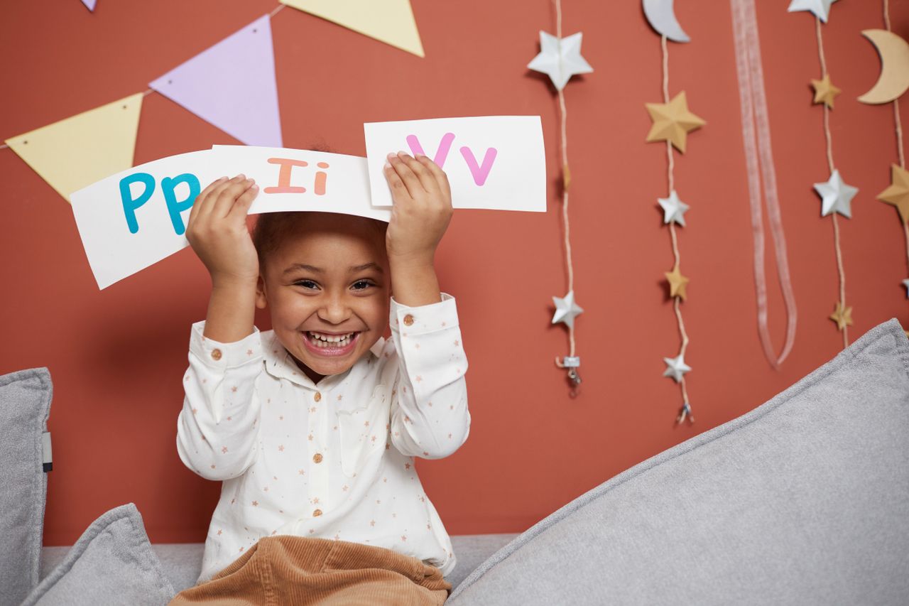 Home Smiling child holding alphabet cards with educational decorations on orange background.